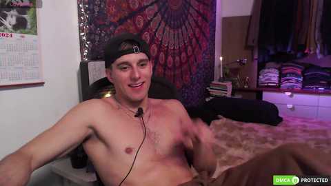 Media: Video of a shirtless, lean, Caucasian man with a black cap, sitting on a bed, smiling. The background features a tapestry, a calendar, a dresser, and a closet.