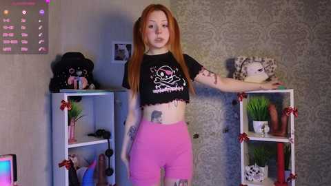 Media: Video of a pale-skinned, red-haired woman with tattoos, wearing a black crop top and pink shorts, leaning against a white shelving unit with red bows. Background includes a plush toy, a green plant, and a framed photo.