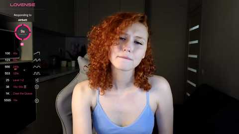 Media: Video of a young woman with curly red hair, wearing a light blue tank top, sitting on a grey chair in a dimly lit kitchen.