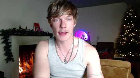 Media: Video of a young man with short, light brown hair, wearing a gray tank top, standing indoors near a lit fireplace and decorated Christmas tree.