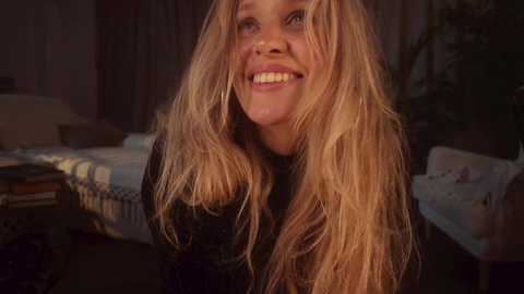 A video of a smiling young woman with long, wavy blonde hair, wearing a black sweater, in a dimly lit bedroom with a bed, nightstand, and wall art.