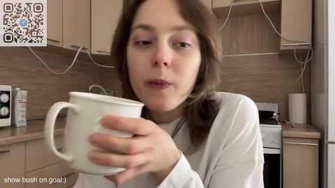 Media: Video of a young woman with brown hair in a white long-sleeve shirt, holding a white mug, in a modern kitchen with wooden cabinets and a stove.