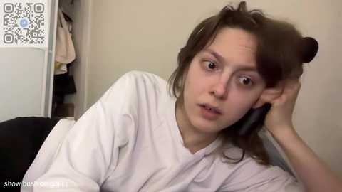 Media: A video of a young woman with light skin and brown hair, wearing a white robe, resting her head on her hand, looking tired and slightly confused. Background shows a cluttered room with a closet and laundry.