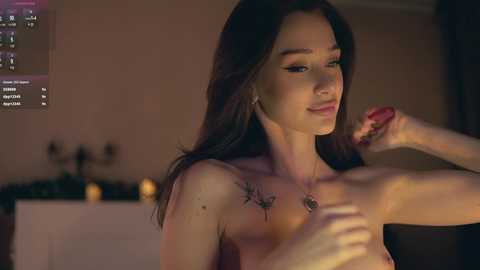 Video of a topless young woman with long dark hair, light skin, and small breasts, applying lotion to her arm in a dimly-lit room.