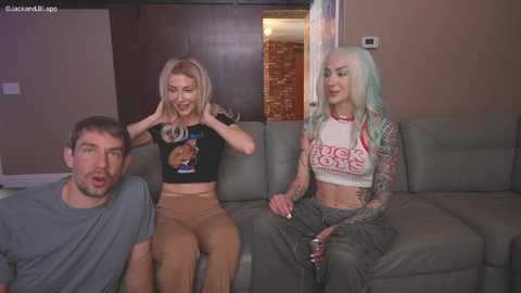 Media: Video of three people: a man, a woman with blonde hair, and a tattooed woman with blue hair, sitting on a gray couch in a living room.