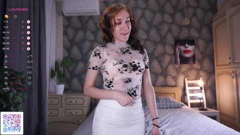 Video of a smiling, fair-skinned, red-haired woman in a sheer black floral top, white skirt, standing in a modern bedroom with a bed, potted plants, and abstract wall art.