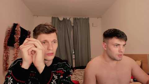 Media: Video of two young, shirtless men in a bedroom. One wears a festive sweater, the other is bare-chested, both looking serious. Background features a bed, curtains, and a tinsel garland.