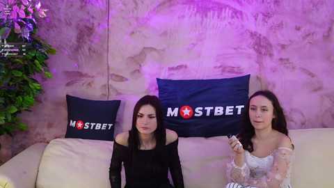 Media: Video of two young women with long black hair, seated on a beige couch, wearing black and white dresses. Background features a textured, beige wall with a \"MSTREET\" pillow and a green plant.