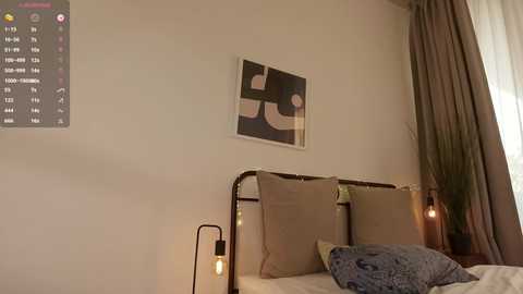 Media: Video of a minimalist bedroom with a metal headboard, beige pillows, a wall clock, abstract art, and a lamp on the bedside table.