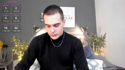 Media: Video of a young, clean-shaven man with short dark hair, wearing a black turtleneck sweater, sitting on a bed with a silver chain necklace. Background shows a grey wall, a lit Christmas tree, and a woman in the background.