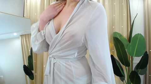 Media: Video of a fair-skinned woman in a sheer, white robe, exposing her ample cleavage. She stands in a well-lit room with beige curtains and a green plant in the background.