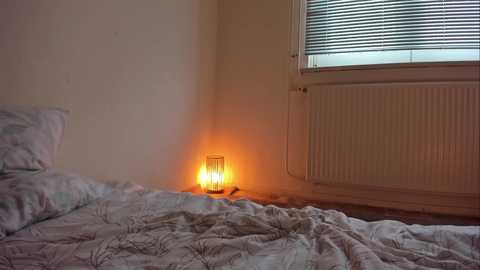 Media: Video of a dimly lit bedroom with a white bed and patterned quilt, a lit candle on a small table, and a window with blinds.