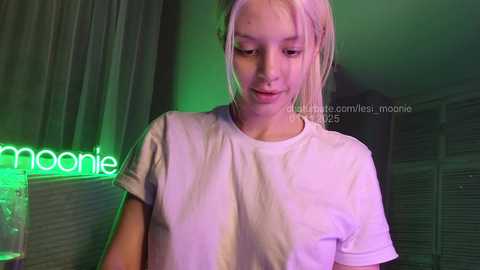 Media: Video of a young, fair-skinned woman with blonde hair, wearing a plain white T-shirt, standing indoors with a green and purple neon light in the background.