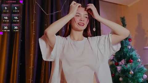 Media: A video of a smiling Asian woman with straight brown hair, wearing a white t-shirt, adjusting her hair in a festive room with a decorated Christmas tree.