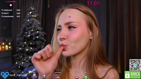 Media: Video of a young, fair-skinned blonde woman with a butterfly tattoo on her forehead, licking her finger seductively in front of a snow-covered Christmas tree and glowing candles in a dimly lit room.