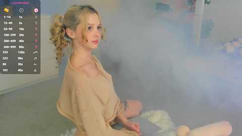 Media: A video of a blonde woman with curly hair, wearing a beige top, sitting cross-legged on a grey floor, surrounded by smoke and blurred background.