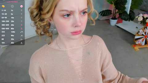 Media: Video of a young, fair-skinned, curly-haired woman with pink lipstick, wearing a light pink sweater, looking pensive, in a modern room with green plants and a digital clock displaying 7:11.