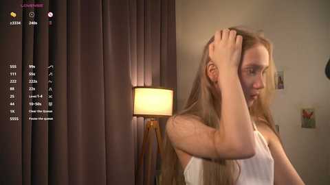 Media: Video of a young woman with long, blonde hair, wearing a white tank top, standing in a dimly lit bedroom with a beige wall, dark curtains, and a lit lamp.