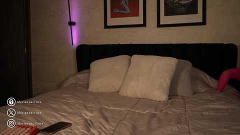 Media: Video of a dimly-lit bedroom with a beige quilted bedspread, two fluffy pillows, and framed artwork on the wall. A smartphone is visible on the bed, and a purple light casts a soft glow.