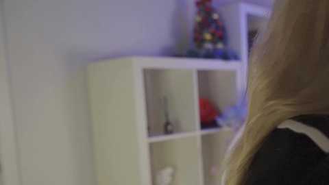 A video of a blonde woman's shoulder, wearing a black shirt, standing beside a white shelving unit with a small, colorful Christmas tree on top. The background is a light-colored room with a slight blur.