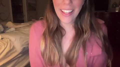 Media: A video of a young woman with long, wavy, light brown hair and fair skin, smiling, wearing a pink sweater. Background shows a dimly lit bedroom with a bed and nightstand.