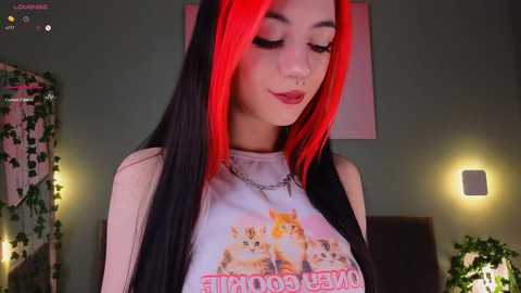 Media: Video of a young woman with long black and red hair, wearing a \"Doom Eternal\" t-shirt, silver jewelry, and a septum piercing. Background includes green walls, fairy lights, and plants.