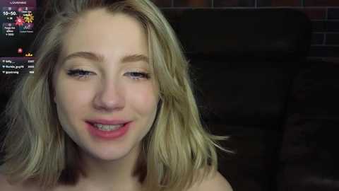 Video of a smiling, light-skinned woman with shoulder-length, wavy blonde hair, wearing minimal makeup, set against a dark background with a brick wall.