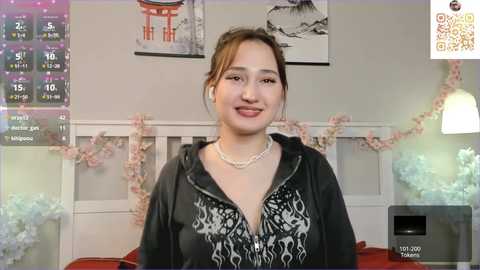 Video of a smiling young woman with light brown skin and dark hair, wearing a black hooded sweatshirt with a white floral pattern, in a cozy, decorated room with pink cherry blossom garlands.