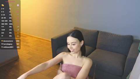 Media: Video of a slim, young woman with light skin and dark hair, sitting on a gray couch in a modern living room, wearing a pink towel.