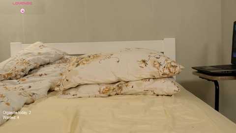 Media: Video of a messy, unmade bed with white sheets and a floral-patterned duvet cover, against a plain beige wall. A black table with a laptop is visible in the background.