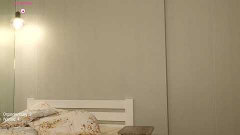 Media: A video of a minimalist bedroom with a white wooden headboard, beige floral bedding, and a beige wall. A single light bulb hangs from the left corner, casting a warm glow.