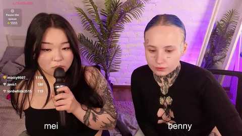 Media: Video of two women with light skin, one with long black hair, tattoos, and green lipstick, and the other with short, slicked-back blue hair, wearing black. They sit in a cozy room with a potted plant and brick wall, using a microphone.