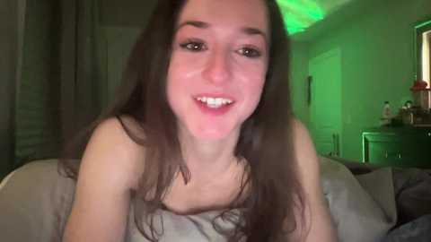 Media: A video of a smiling, slender, young woman with long dark hair, fair skin, and no visible clothing, lying on a bed in a dimly lit room with green LED lights.