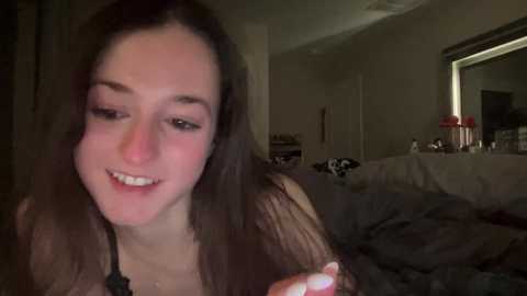 Media: A dimly lit video of a young woman with straight, brown hair, smiling, in a messy bedroom. She has fair skin and is wearing a black top.