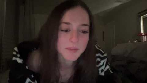 Media: Video of a young woman with long, straight brown hair, wearing a black and white patterned top, sitting in a dimly lit bedroom, with a blurred background featuring a bed and a window.