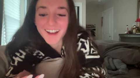 Media: A video of a young woman with long dark hair, smiling, wearing a black and white patterned sweater, sitting on a bed with messy gray sheets. Background shows a bedroom with a door, dresser, and a window with blinds.