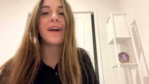 Media: Video of a young woman with long, straight, light brown hair, wearing a black shirt and black jacket, smiling with parted lips, in a white room with a tall white cabinet and a purple decorative item.