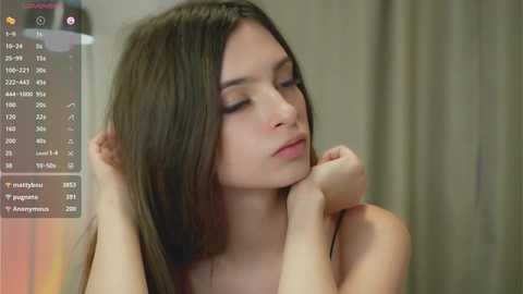 Video of a young woman with long, straight brown hair, wearing a soft expression, touching her face, in a neutral-colored room.