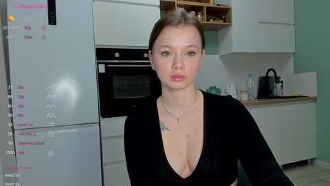 Media: Video of a young woman with fair skin and brown hair, wearing a black V-neck top, standing in a modern kitchen with white cabinets, a microwave, and a calendar on the fridge.