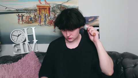 Media: Video of a young man with straight black hair, wearing a black T-shirt, brushing his hair on a gray leather couch. Background includes a colorful wall mural, a white clock, and a fluffy pink pillow.