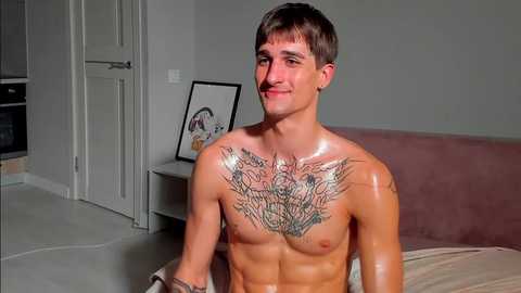 Media: Video of a shirtless, fit young man with a tattoo of a lion's head across his chest, sitting on a bed in a modern, gray-toned room with a framed art piece on a white shelf.