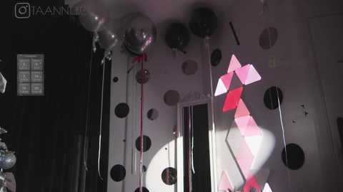 Media: Video of a dimly lit room with white walls adorned with black polka dots and a pink geometric pattern. Balloons and string lights hang from the ceiling.