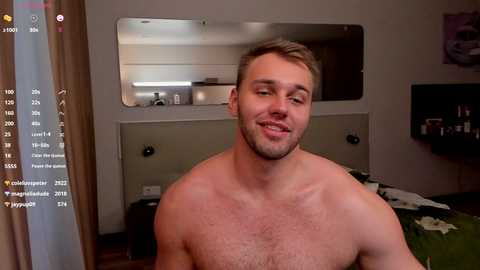 Media: Video of a shirtless, muscular, fair-skinned man with short blond hair, smiling, standing in a dimly lit bedroom with a green bed, beige curtains, and a mirror on the wall.