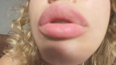 Media: A close-up video of a woman's full, slightly parted lips, with light pink lipstick, set against a blurred background of wavy, blonde hair and a brown couch.