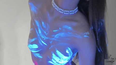 Media: Video of a topless woman with long, dark hair, covered in blue body paint. She wears a sparkling necklace, creating a contrast with her smooth, shiny skin. Background is a plain, light-colored wall.