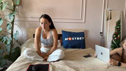 Media: Video of a young woman in white pajamas meditating on a bed with a laptop and Christmas decorations, surrounded by plants and a blue pillow.