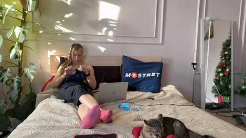 Media: Video of a blonde woman in a dark blue tank top and gray shorts, seated on a bed with a gray cat, laptop, and a blue \"MOSTREET\" pillow in a sunlit room.