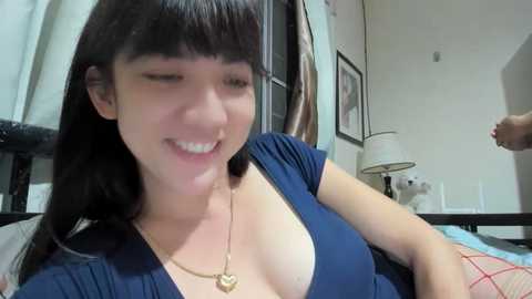 Media: Video of a young East Asian woman with straight black hair, wearing a low-cut blue top, smiling, in a bedroom with a lamp and framed artwork.