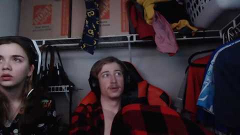 Media: A video of a young man with headphones on, wearing a red and black plaid shirt, sitting in a messy closet with various clothes and boxes in the background.