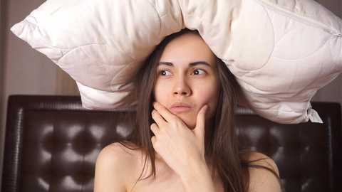 Media: Video of a young woman with long brown hair, covered by a large, white, quilted pillow, looking contemplative. She has fair skin and is topless. Background shows a dark brown leather couch.